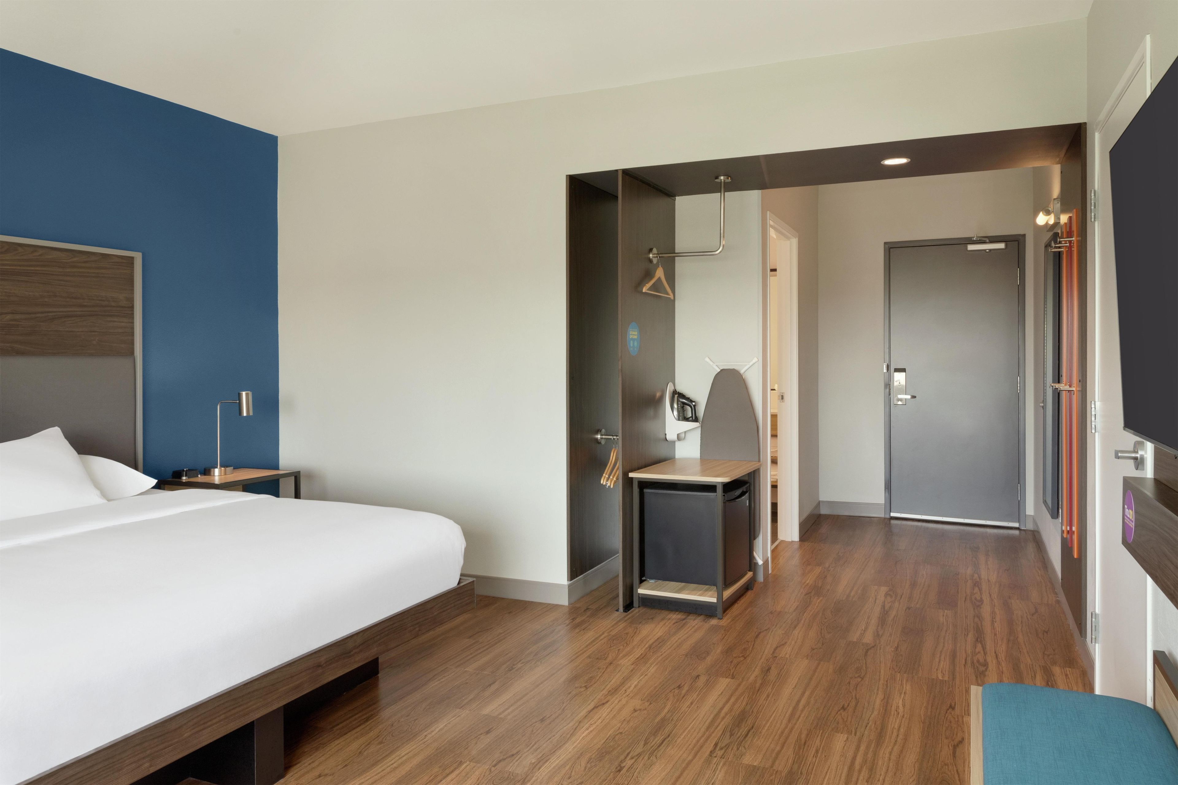 Habitación de hotel con cama, mesita de noche y un pequeño escritorio con silla. Se observa una puerta que da a otra habitación y un perchero.