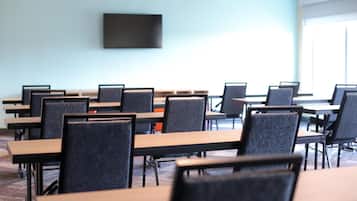 Une salle de conférence avec des rangées de tables et de chaises, un téléviseur à écran plat fixé au mur et de grandes fenêtres laissant entrer la lumière naturelle.