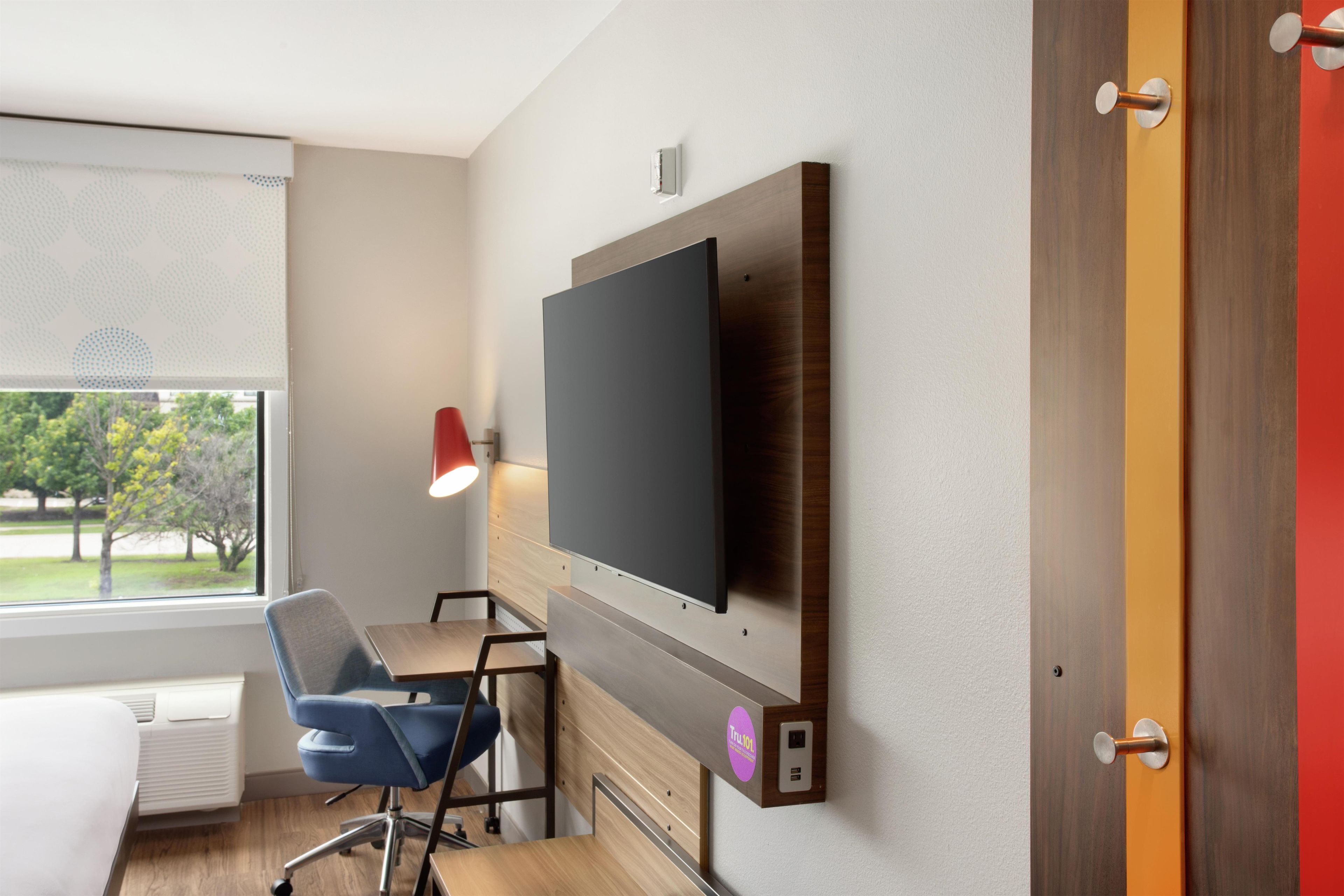 Une chambre d’hôtel moderne équipée d’un téléviseur à écran plat fixé au mur, d’un bureau avec une chaise, d’une fenêtre avec des stores et d’une porte dotée d’un cadre rouge.