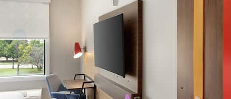 A modern hotel room with a flat-screen TV mounted on the wall, a desk with a chair, a window with blinds, and a door with a red frame.