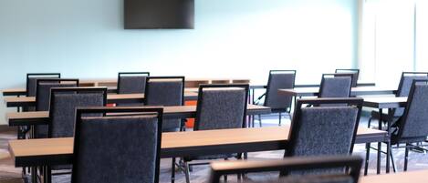 A conference room with rows of tables and chairs, a flat-screen TV mounted on the wall, and large windows.