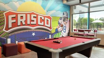Una mesa de billar con superficie de fieltro rojo, un mural con la palabra 'FRISCO' y un jugador de fútbol, y vistas a un parque a través de amplias ventanas.