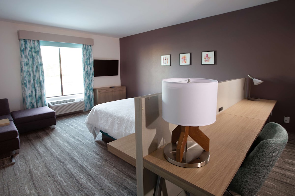 A hotel room with a bed, a desk with a lamp, a chair, a TV mounted on the wall, and a window with curtains.