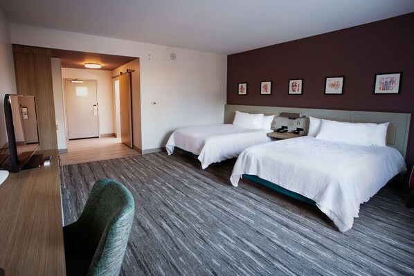 A hotel room with two beds, a desk, and a chair. There is a door leading to another room and a wall with framed pictures. - Hilton Garden Inn Southern Pines Pinehurst, NC (Aberdeen)