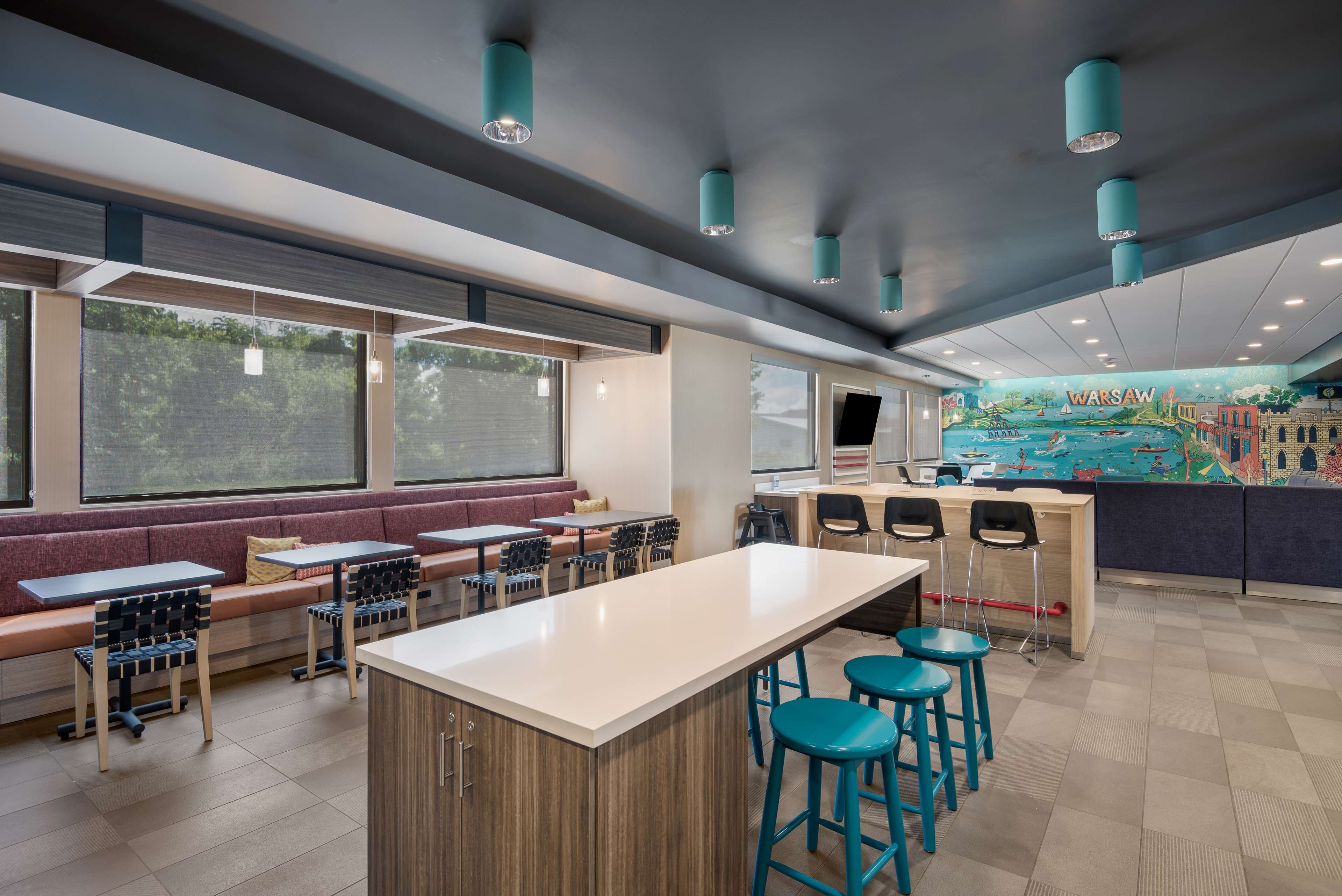 A modern restaurant with a central bar area, seating along the windows, and a mural of Warsaw on the back wall.