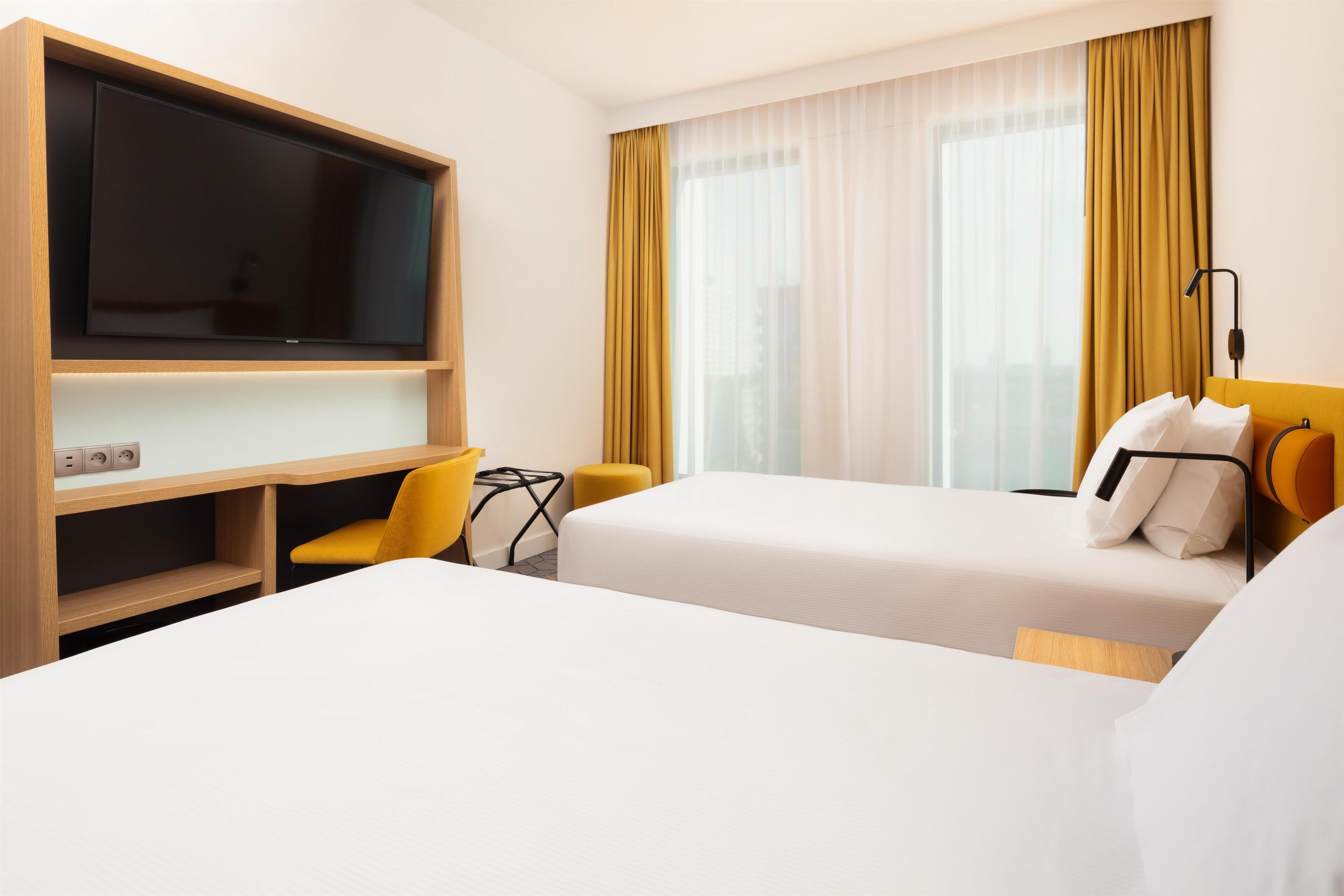 Una habitación de hotel con cama, escritorio con televisor, una silla y ventana con cortinas.