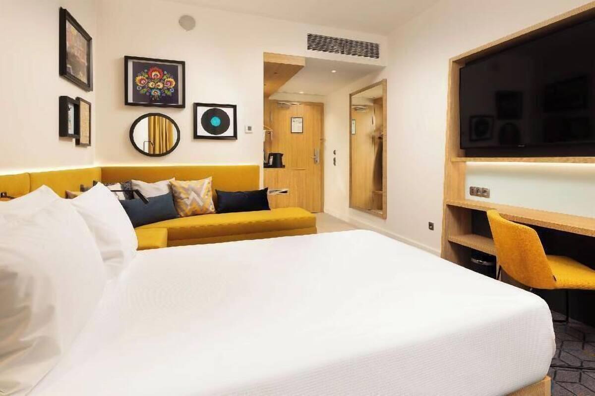 Quarto de hotel com cama, sofá amarelo, escrivaninha e espelho.