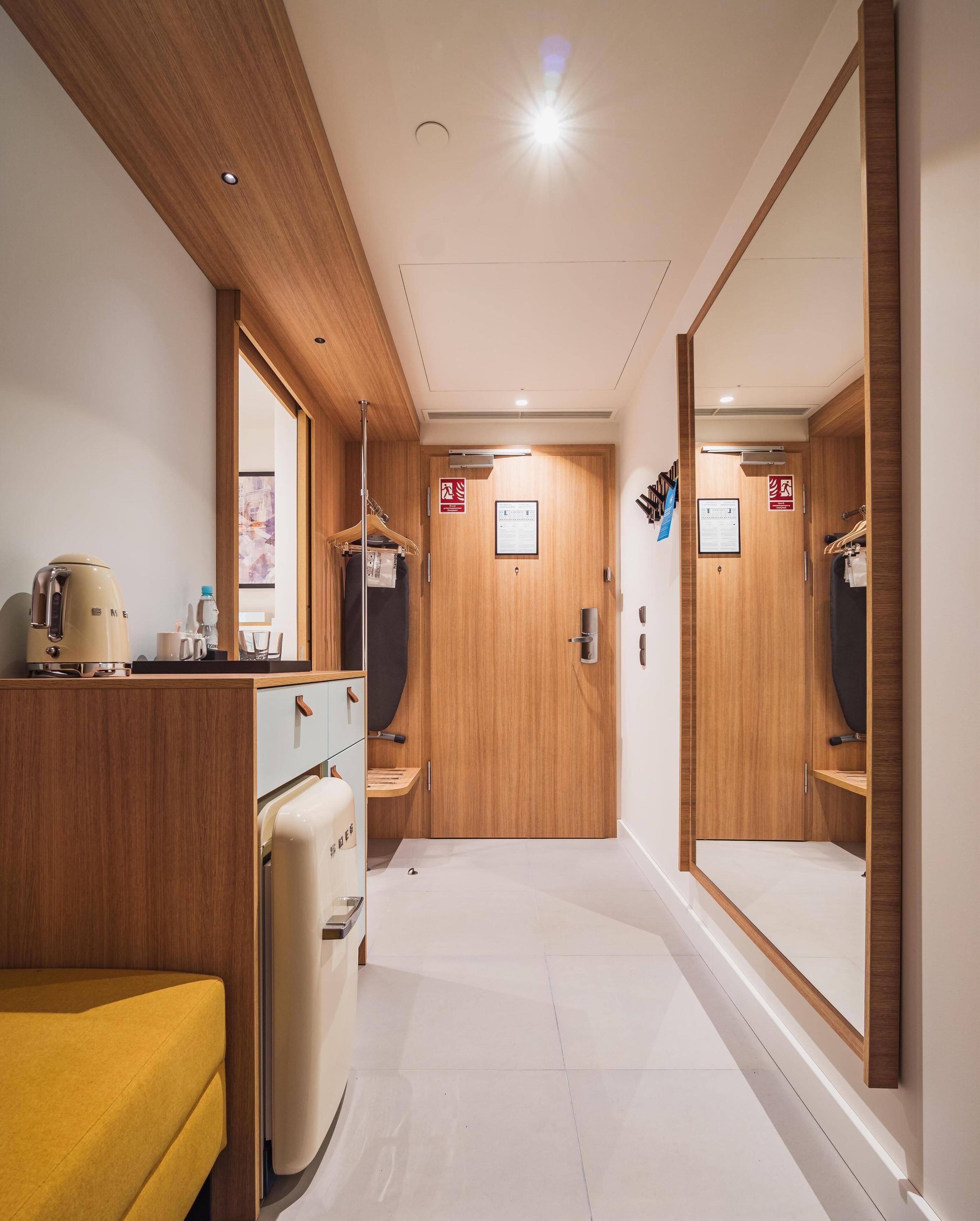 A hotel room with a wooden wardrobe, a kitchenette, a yellow sofa, and a mirror.