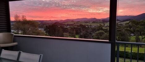A balcony with a table and chairs, overlooking a landscape with trees and distant hills during sunset.