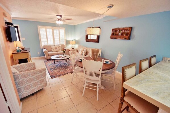 A living room with a sofa, a dining table with chairs, a ceiling fan, and a TV mounted on the wall. - Demere House Condo #2 | Walk-to-Beach 2BR St. Simons Island Vacation Rental Near Pier Village (St. Simons)