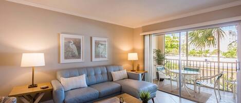 A modern living room with a grey sofa, two side tables with lamps, a wooden coffee table, and a balcony with outdoor furniture.