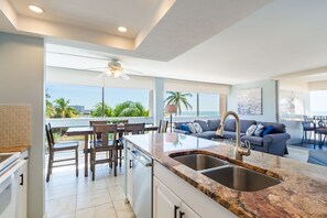 A modern kitchen with a large island, a dining area with wooden chairs, and a living area with a sofa and a view of the ocean.