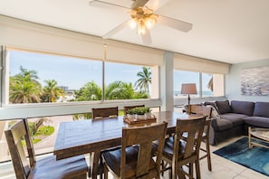 A spacious living area with a large dining table, comfortable seating, and a view of palm trees and a beach.