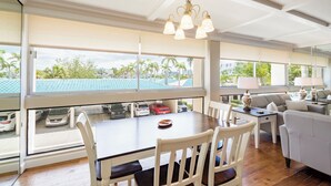 Amplia habitación con una gran mesa de comedor, sillas blancas, una araña y vistas al exterior con coches estacionados y palmeras.