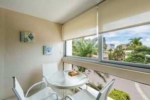 Un balcón con una mesa de vidrio, sillas blancas y vistas a palmeras y edificios.