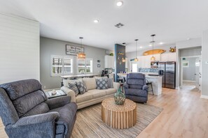 A modern living room with a sofa, armchairs, and a coffee table. A kitchen with a dining area is visible in the background.