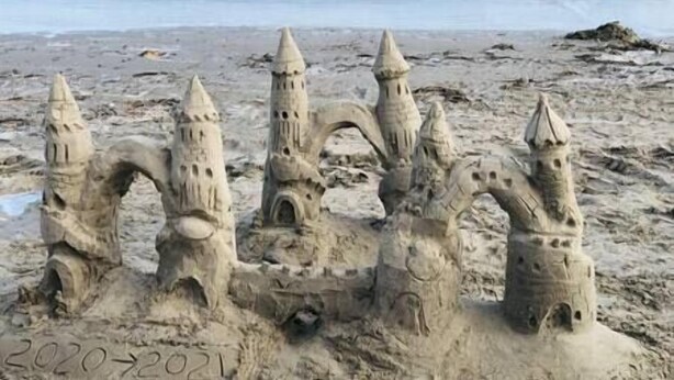 Ein Sandburg mit mehreren Türmen und Bögen, auf einem sandigen Strand mit Wellen im Hintergrund.