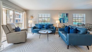 Uma sala de estar moderna com um sofá bege, um sofá azul e uma mesa de centro redonda. Há duas janelas com persianas, um quadro na parede e uma luminária em cada lado do quarto.