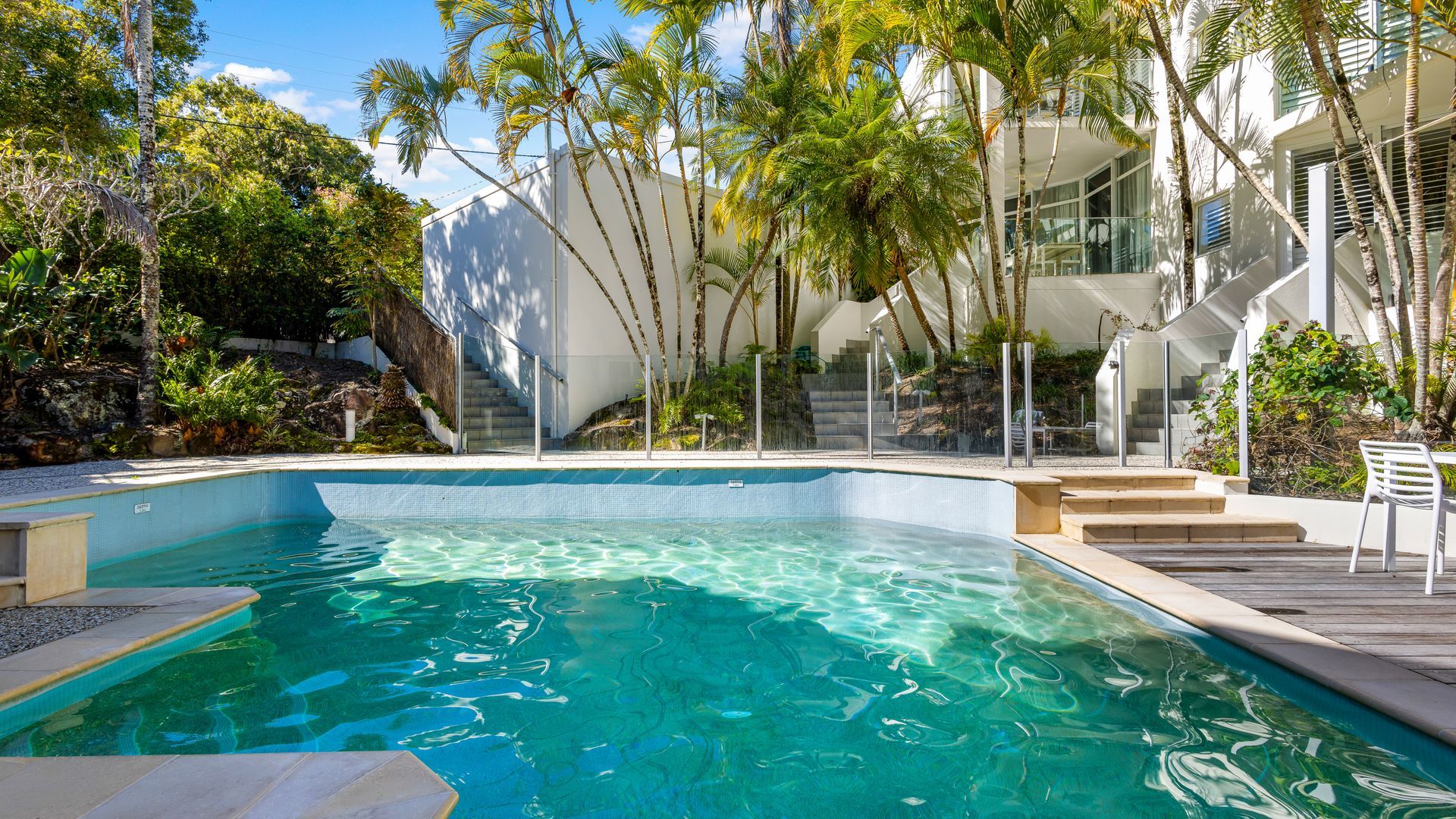 A swimming pool with clear water, surrounded by palm trees and a modern building with a staircase leading up to a balcony.