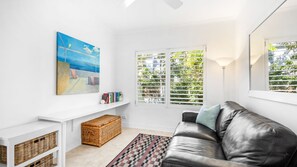 A modern living room with a leather sofa, a painting, a bookshelf, a wicker basket, and a window with blinds.