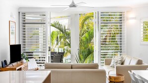A modern living room with a sofa, a coffee table, and a TV. There are large windows with blinds, and a ceiling fan is visible. - Upper Hastings Penthouse 5 Noosa Heads (Noosa Heads)