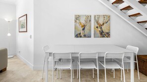 Uma sala de jantar com uma mesa e cadeiras brancas, uma escada e duas pinturas de veados penduradas na parede.
