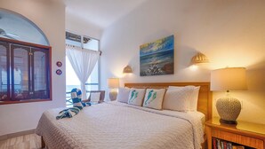 A bedroom with a large bed, a window with curtains, a painting on the wall, and a nightstand with books. - Beachfront Penthouse Condo with Two Oceanview King Suites (Akumal)