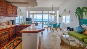 Een ruime keuken met een groot eiland, een eetgedeelte met uitzicht op zee, en een gezellige zithoek met een groene bank.