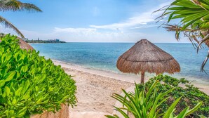 A beach with a thatched umbrella, clear blue water, and lush greenery. - Newly Updated Beachfront Condo with Terrace, Pool & Daily Service (Akumal)