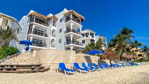 A beachfront hotel with multiple balconies, a stone staircase leading to the beach, and blue lounge chairs. - Newly Updated Beachfront Condo with Terrace, Pool & Daily Service (Akumal)