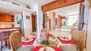 A spacious living area with a glass-top dining table surrounded by wicker chairs, a large decorative urn, and a view into the adjacent bedroom. - Oceanview Condo with Balconies, Pool & Daily Service (Akumal)