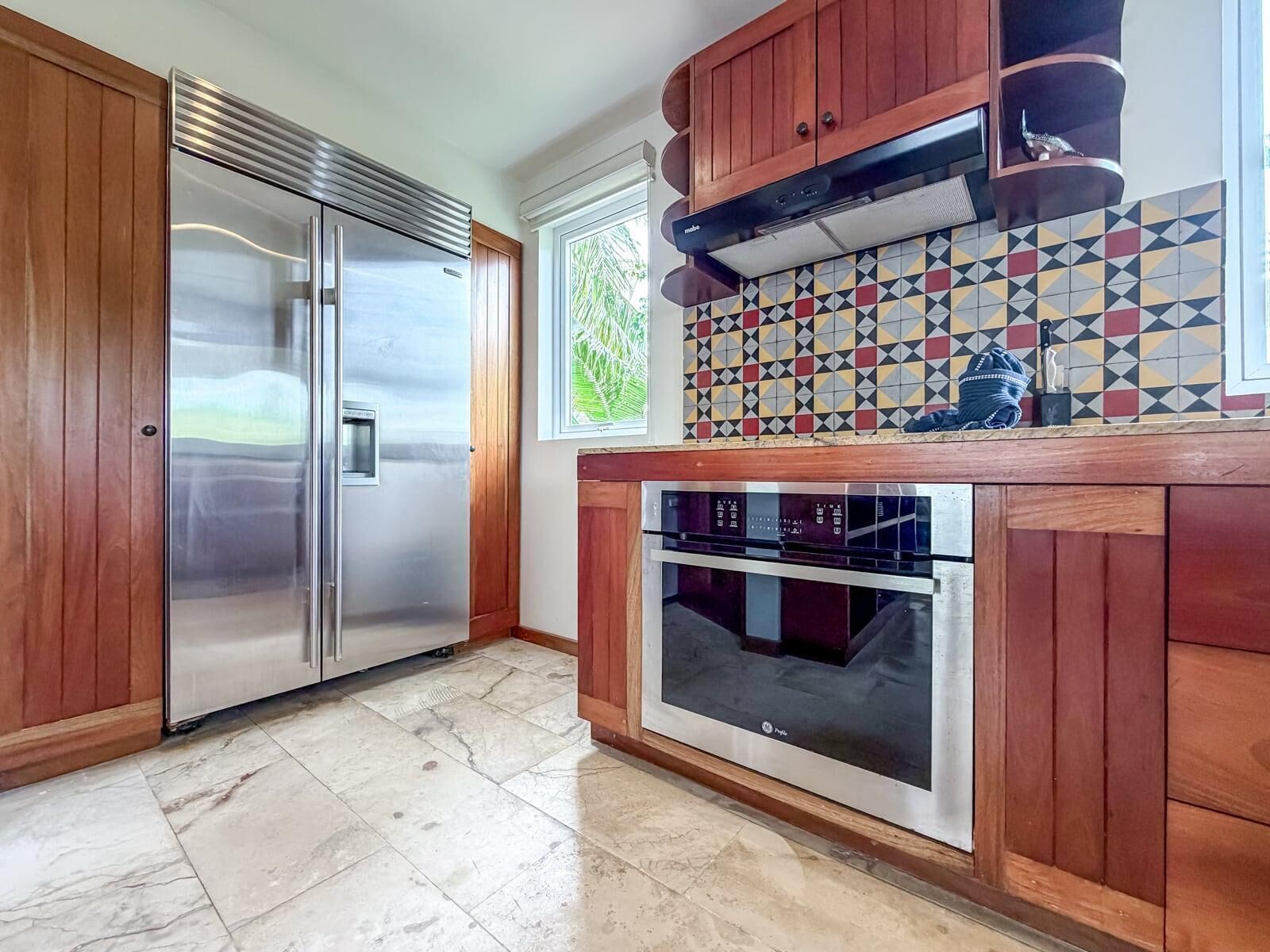 A kitchen with a stainless steel refrigerator, a built-in oven, wooden cabinets, and a patterned backsplash.