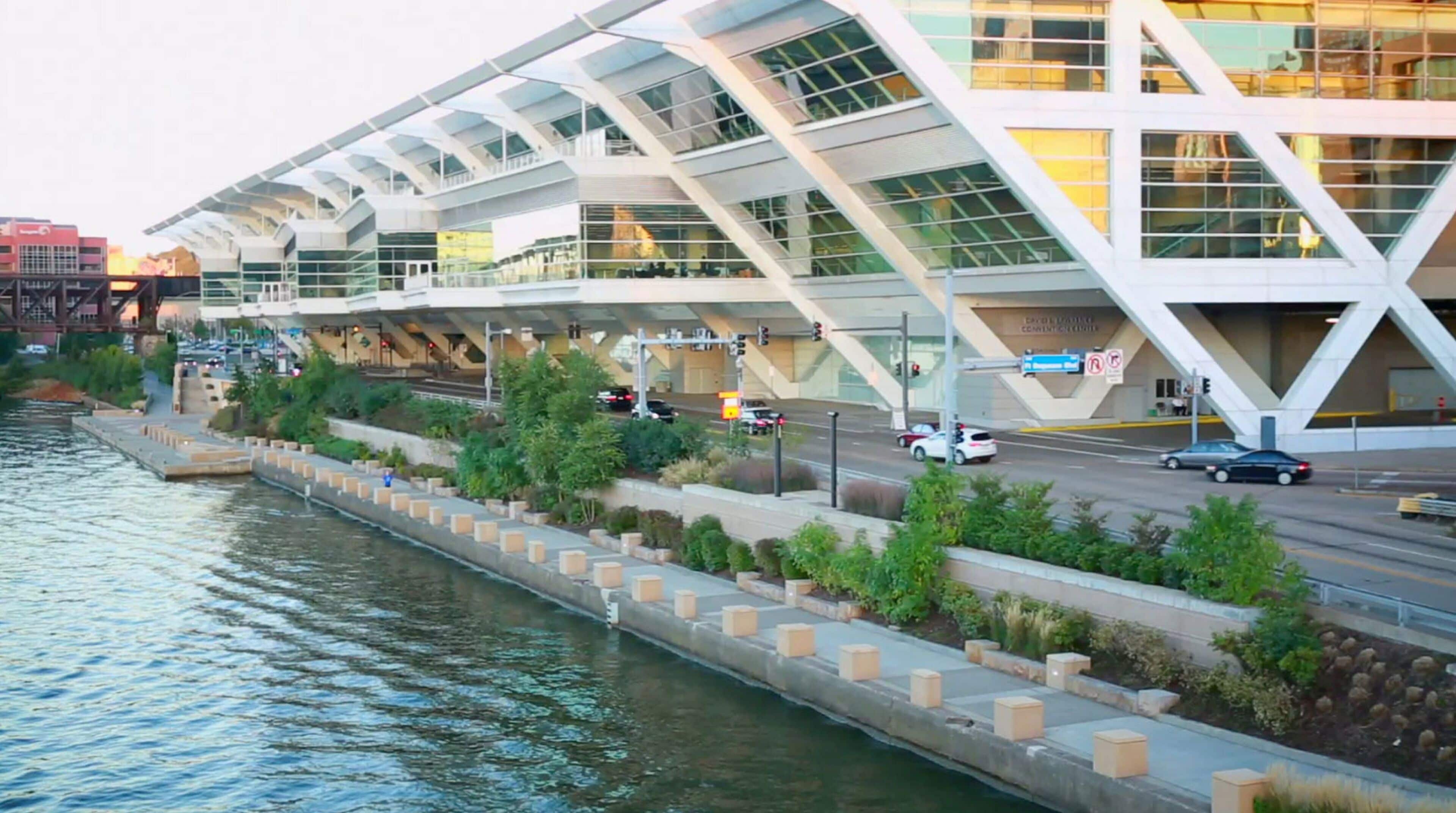 A modern building with a unique angular design, located beside a river with a pedestrian walkway and greenery.
