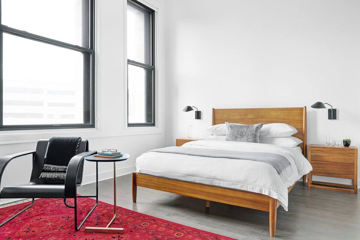 A modern bedroom with a wooden bed, two bedside tables with lamps, a black chair, a small side table with a book and glasses, and a red patterned rug.