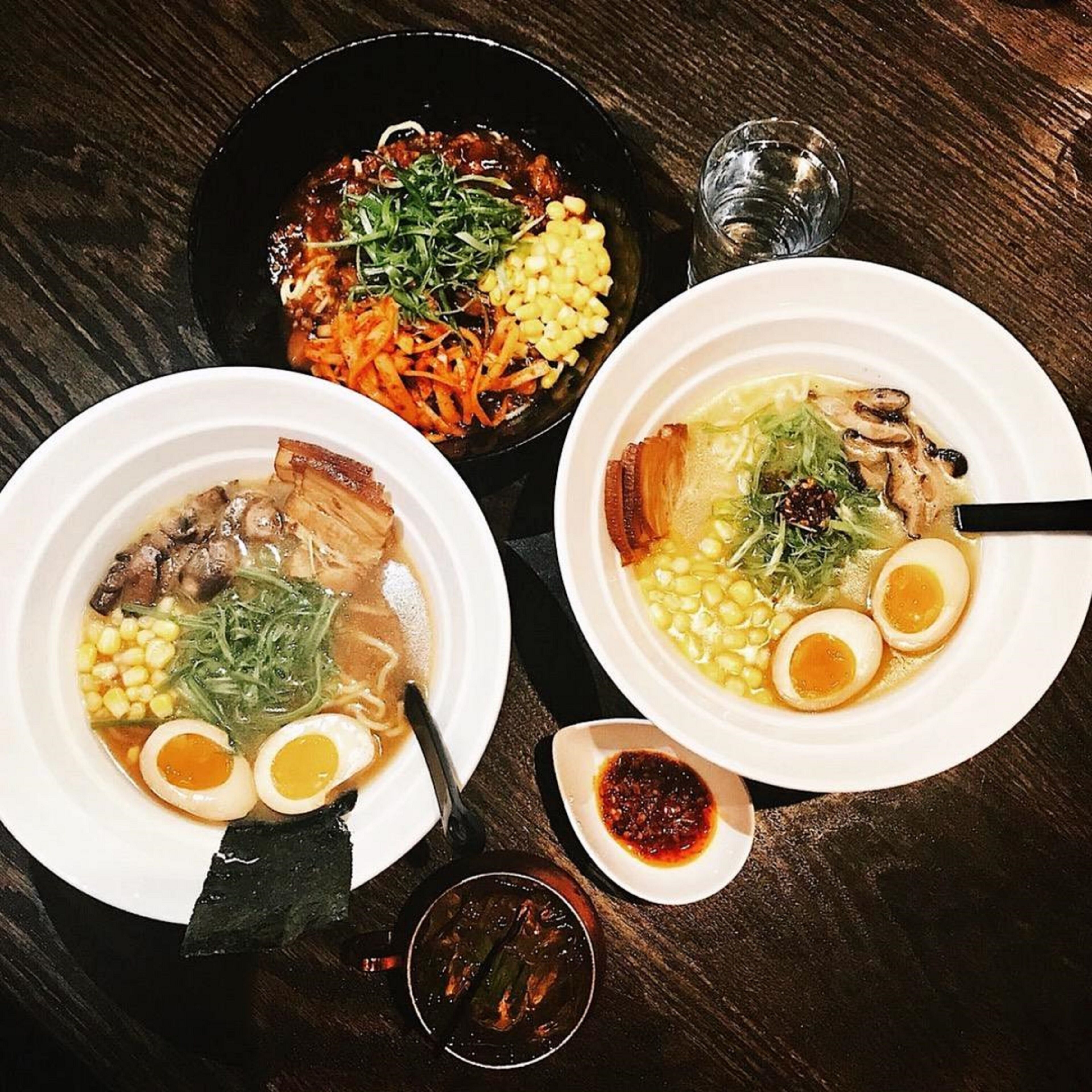 Three bowls of soup with various toppings, a small dish of sauce, and a glass of water on a wooden table.