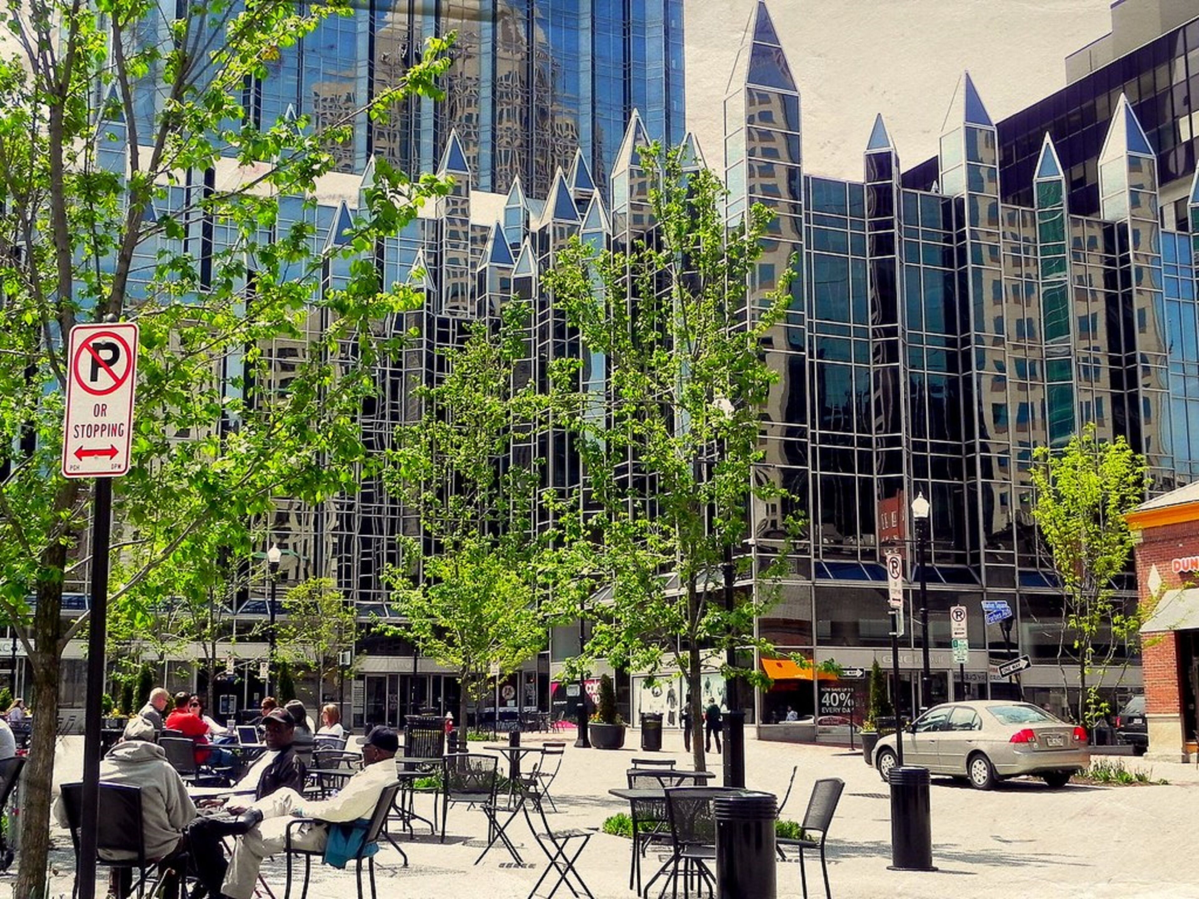 Un espacio al aire libre con mesas y sillas, una señal de prohibido estacionar y un edificio moderno con ventanas de vidrio reflectante al fondo.