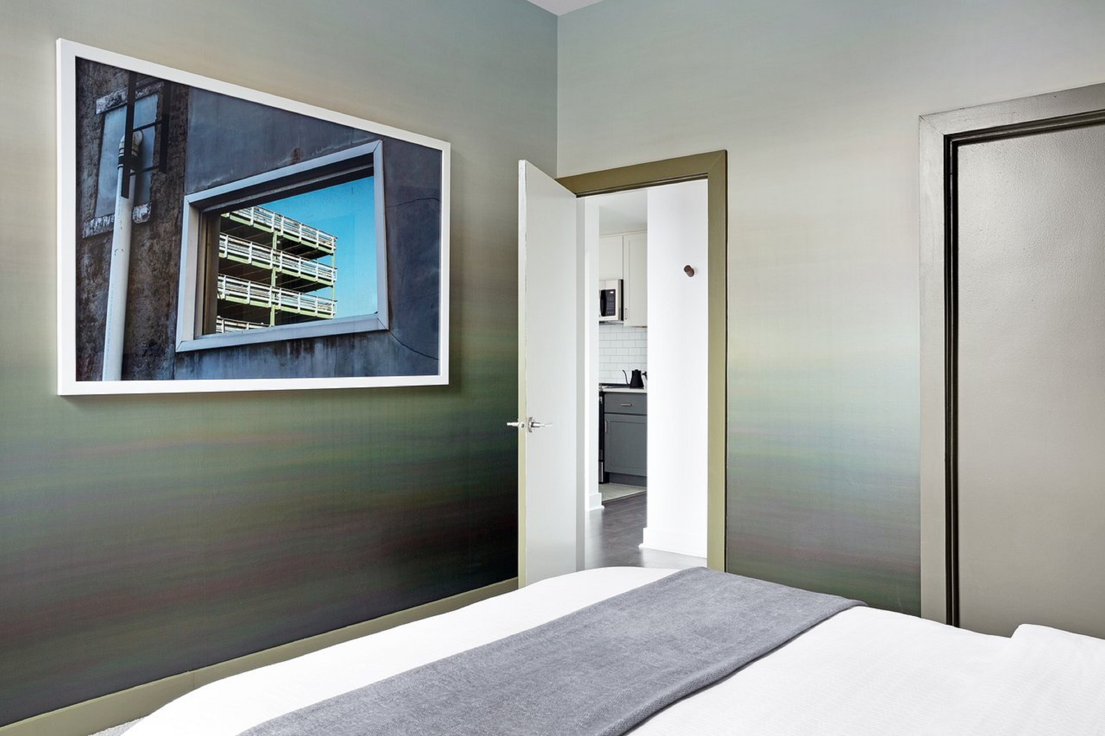Una camera d'albergo moderna con un letto, un'ampia foto incorniciata di un edificio e vista sull'angolo cottura.