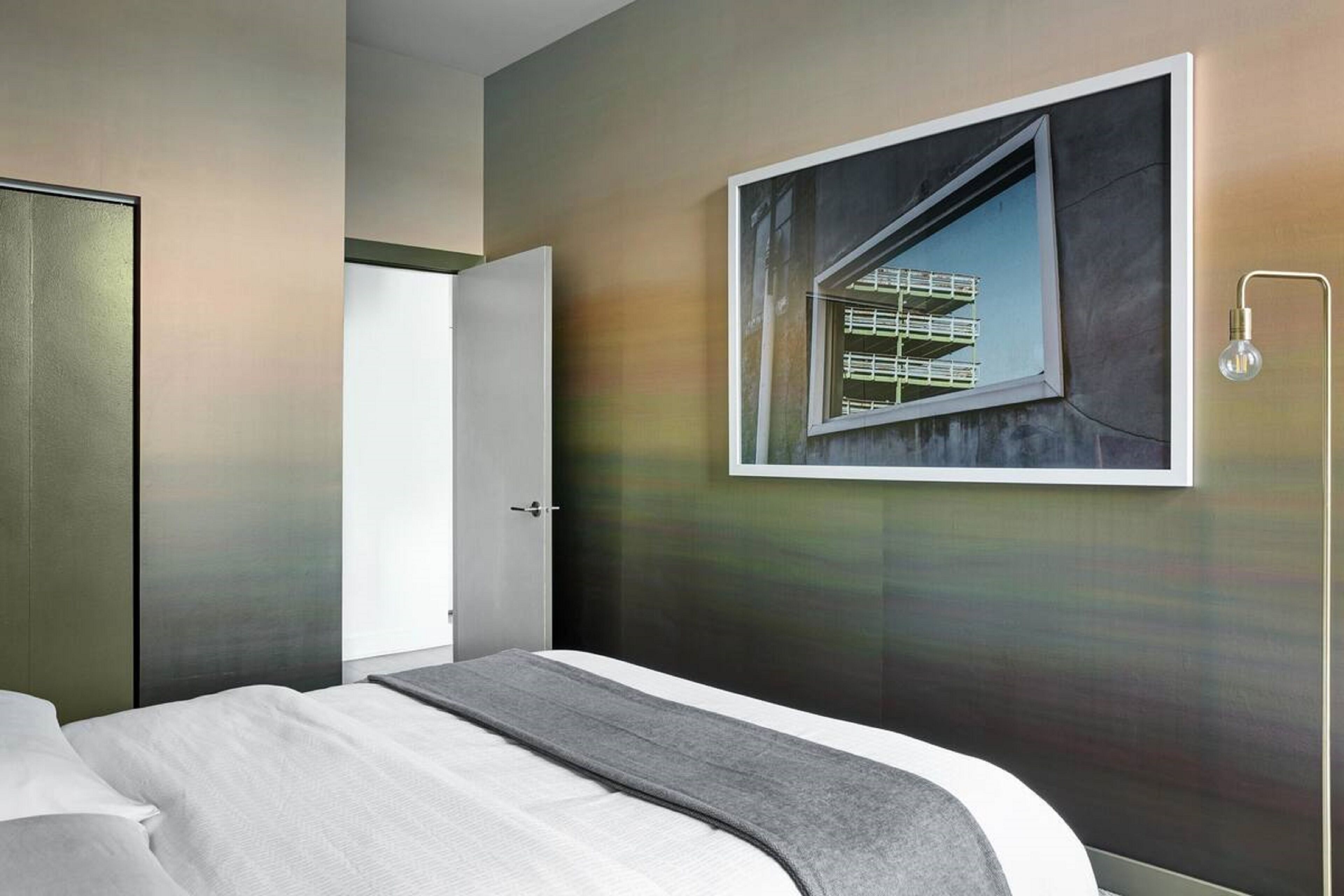 A modern hotel room with a bed, a door, and a large framed picture of a building.