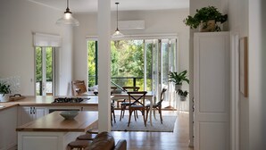 A spacious kitchen with a central island, wooden dining table, and chairs. There are large windows providing natural light and views of greenery outside. - Pembroke Cottage- modern comforts and scenic views (Daylesford)