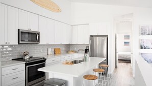 Een moderne keuken met witte kasten, roestvrijstalen apparatuur, een groot eiland met krukken, en een uitzicht op een slaapkamer door een geopende deur.