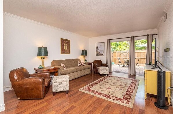 A living room with a sofa, two armchairs, a coffee table, and a rug. There is a sliding glass door leading to an outdoor area with a wooden fence.