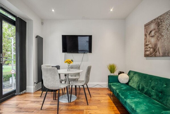 A modern living room with a green sofa, a round dining table with four chairs, and a flat-screen TV mounted on the wall.