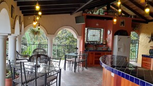 Un área de comedor al aire libre techada con mesas y sillas, una barra de cocina y lámparas colgantes.