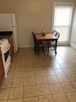 A kitchen with a stove, oven, and refrigerator, a wooden dining table with chairs, and a window with blinds. - 1 bedroom/1 bath (Oneonta)