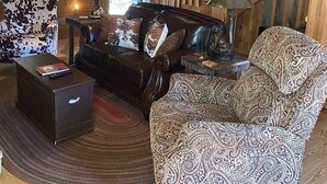 Una sala de estar acogedora con una escalera de madera, un sofá de cuero, un sillón estampado y una mesita de noche con una lámpara.