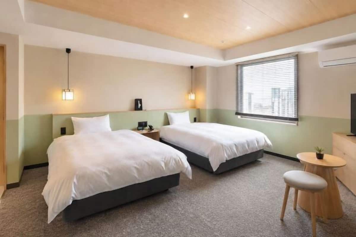 Ett hotellrum med två sängar, ett skrivbord, en stol och ett fönster med persienner.