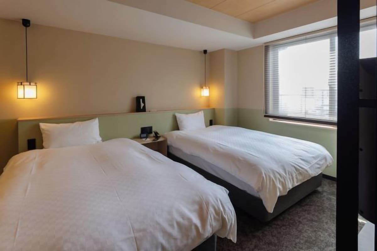 Habitación de hotel con dos camas individuales, una mesita de noche, una lámpara y una ventana con persianas.
