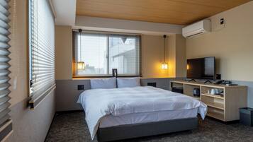 Une chambre d’hôtel avec un lit, un bureau avec un ordinateur, une télévision et une fenêtre avec des stores.