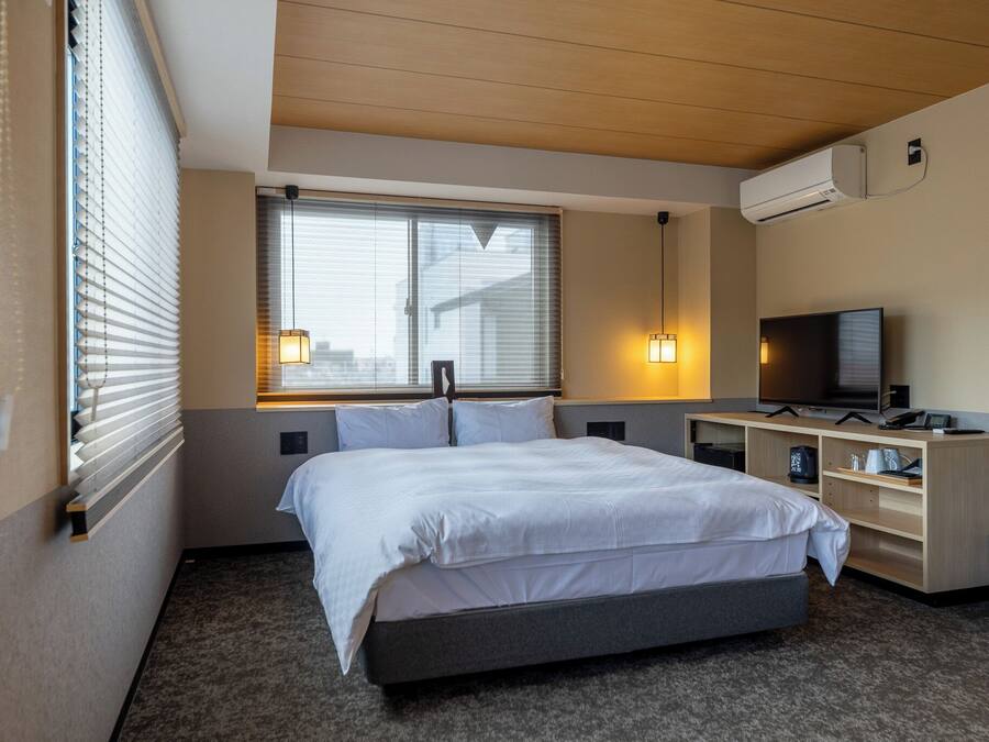 Une chambre d’hôtel avec un lit, un bureau avec un ordinateur, une télévision et une fenêtre avec des stores.