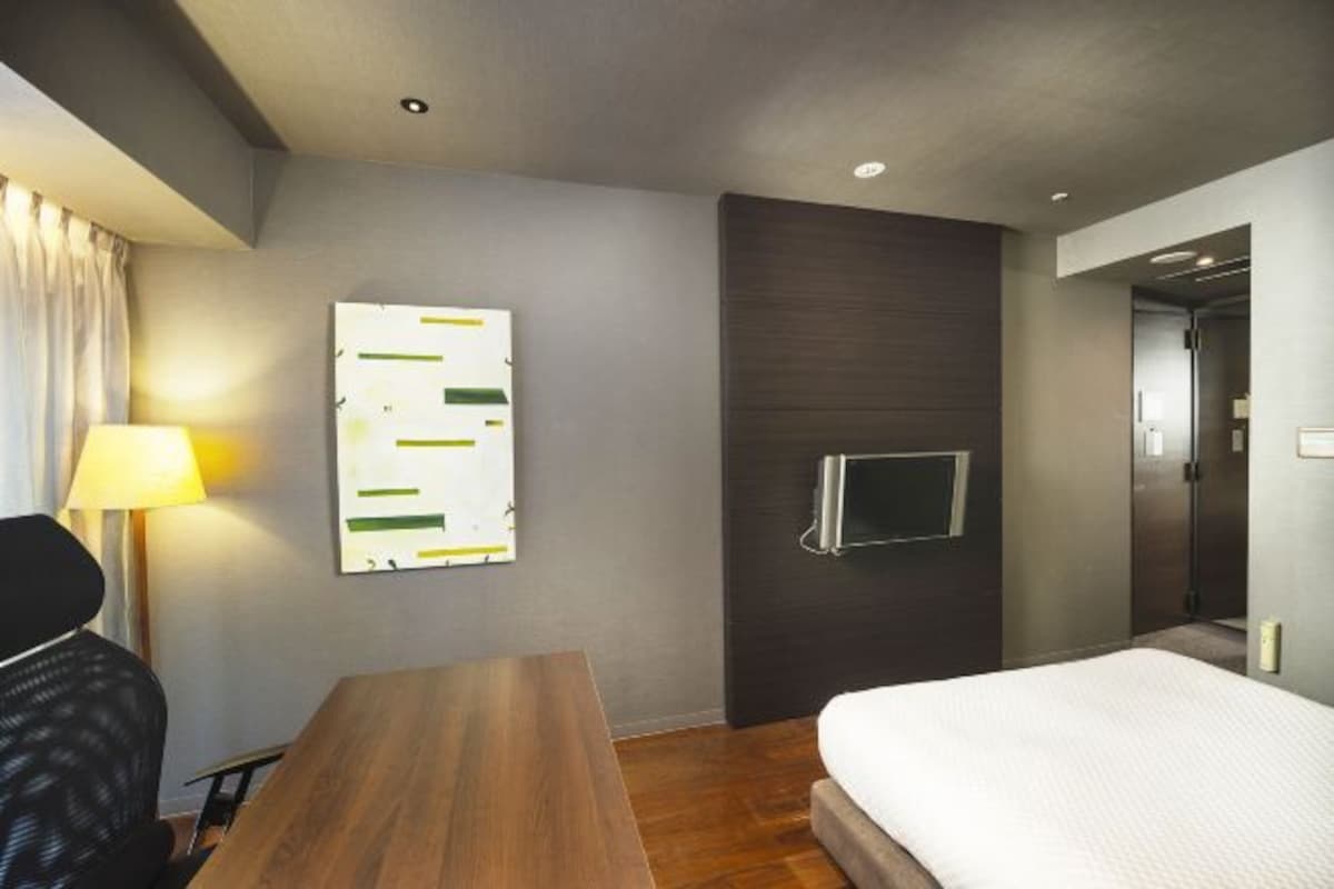 Una habitación de hotel con un escritorio de madera, una cama, un televisor montado en la pared y un cuadro.